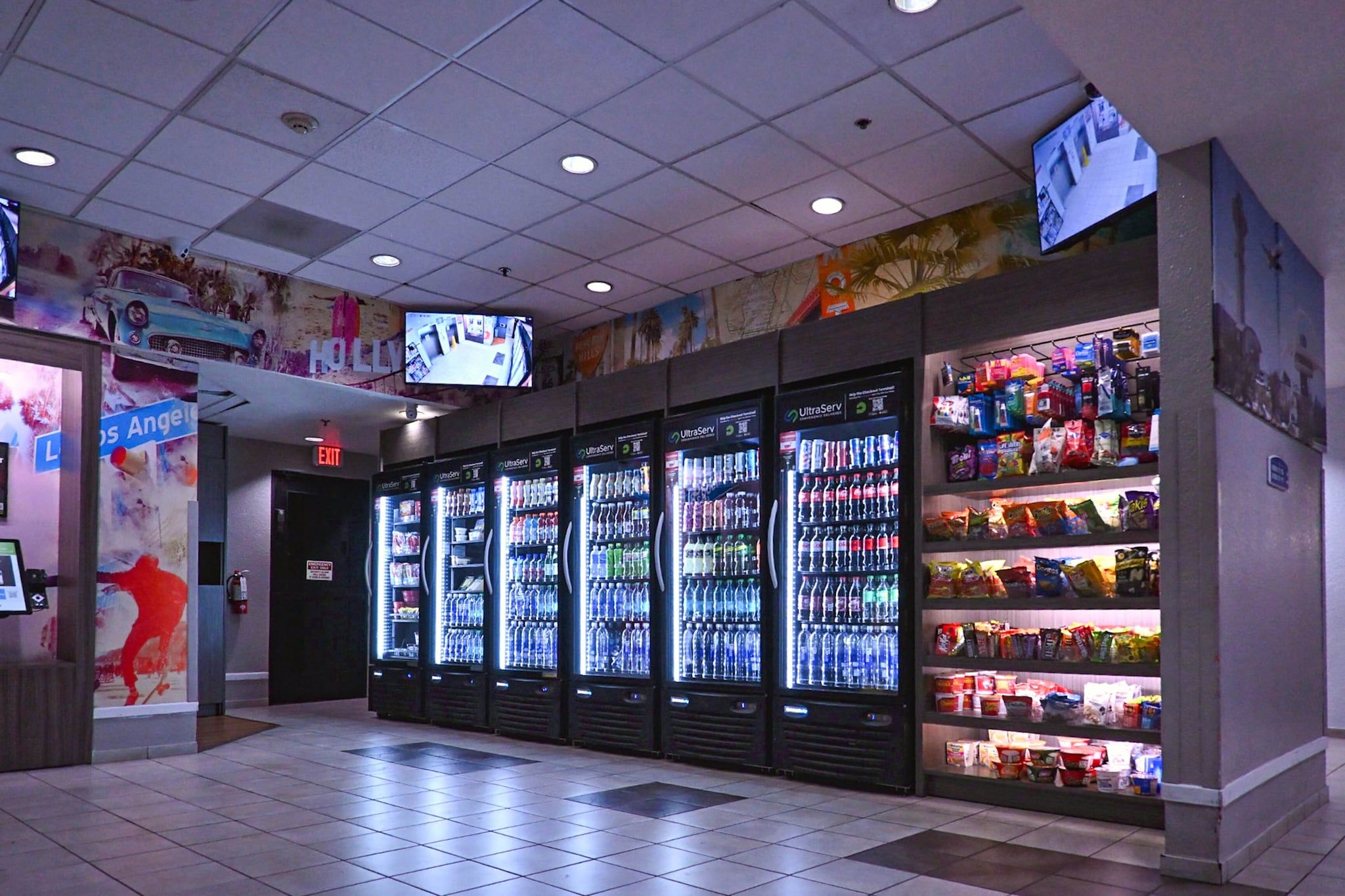A convenience store with multiple beverage dispensers, a selection of snacks, and a television mounted on the ceiling.