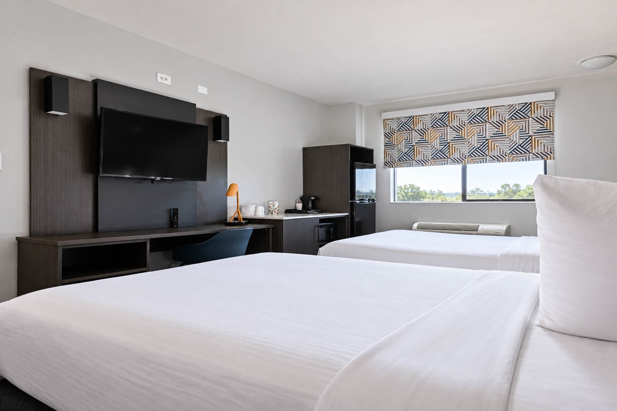 Een hotelkamer met twee bedden, een bureau met een computer, een televisie en een raam met een decoratief zonnescherm.