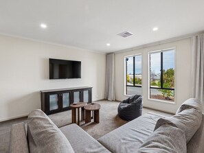 Ein modernes Wohnzimmer mit Fernseher, einem dunklen Schrank, einem grauen Sofa, einem Sitzsack und Blick ins Freie durch große Fenster.
