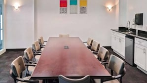 Un salón de conferencias con una larga mesa, sillas y una zona de cocina con armarios y un microondas.