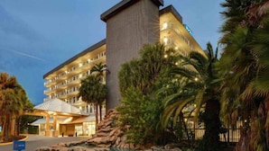 Un hotel de varios pisos con techo plano, entrada cubierta y zona ajardinada con palmeras y rocas.