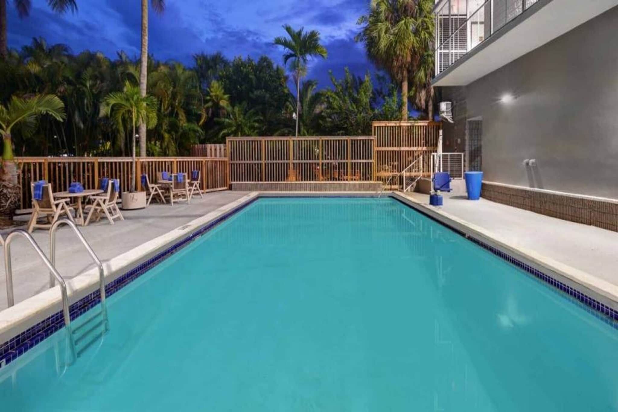 A swimming pool with a wooden deck, lounge chairs, and a view of palm trees.