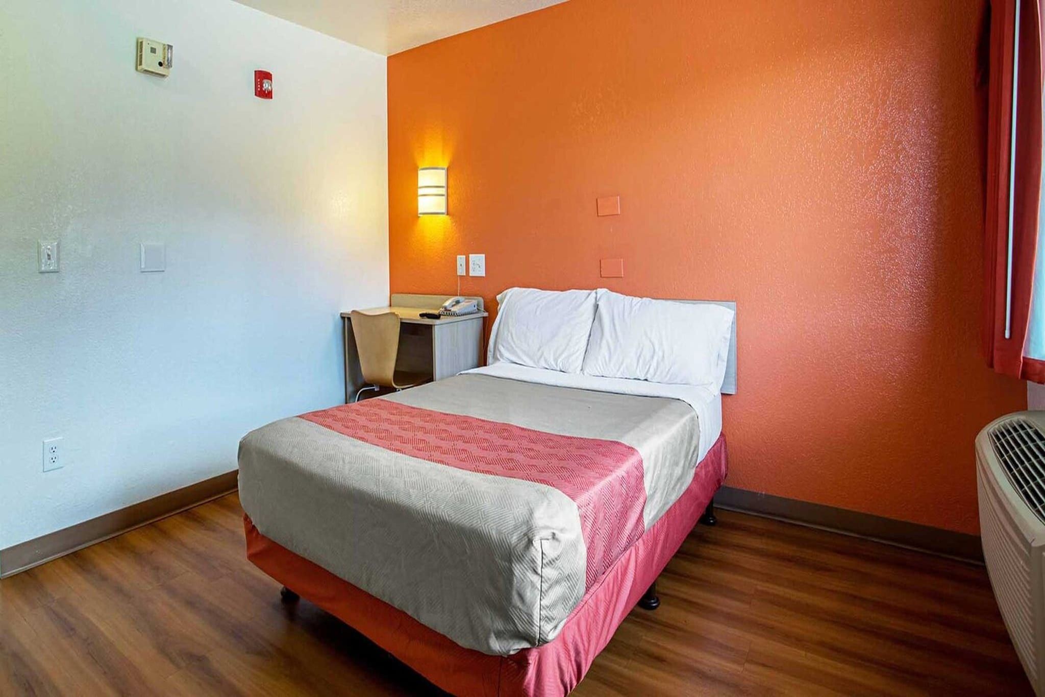 Une chambre d’hôtel comprenant un lit, un bureau, une chaise, une table de chevet, un radiateur fixé au mur et une fenêtre avec des rideaux.