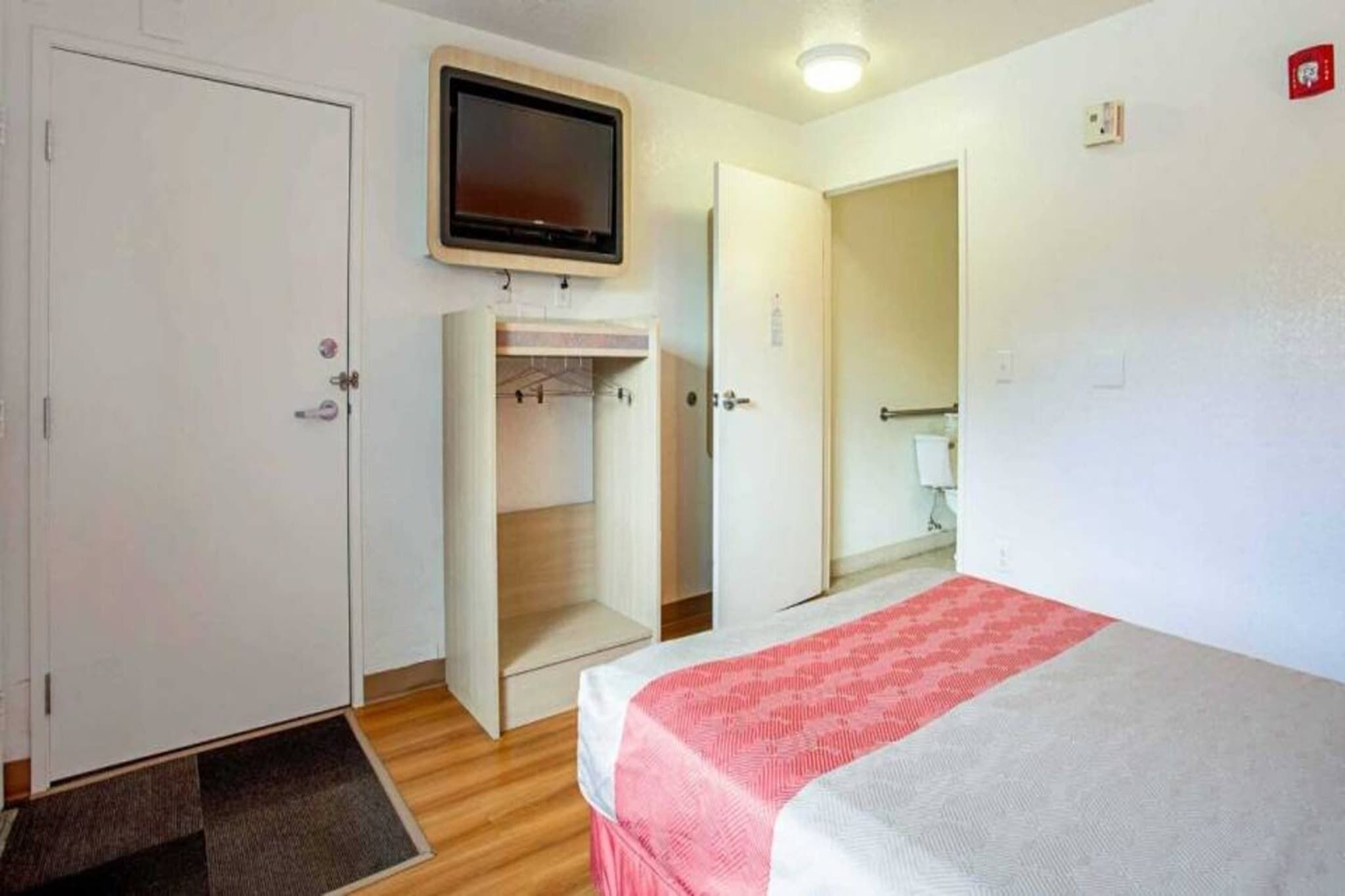 Ein Schlafzimmer mit einem Bett, einem an der Wand montierten Fernseher, einem Kleiderschrank und einem durchgehenden Türbogen einsehbarer Badezimmer.