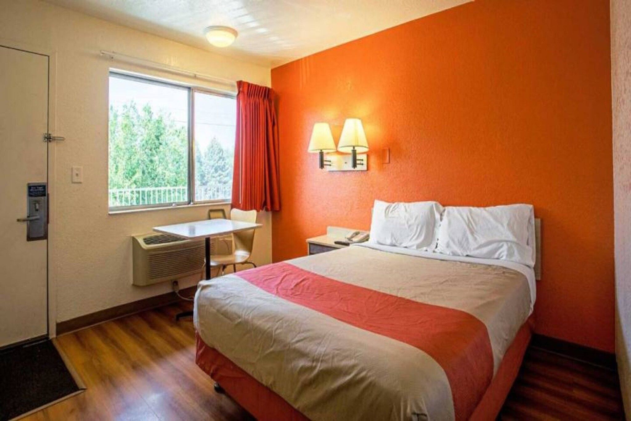 Une chambre d’hôtel comprenant un lit, un bureau, une chaise, une fenêtre avec des rideaux et une lampe fixée au mur.