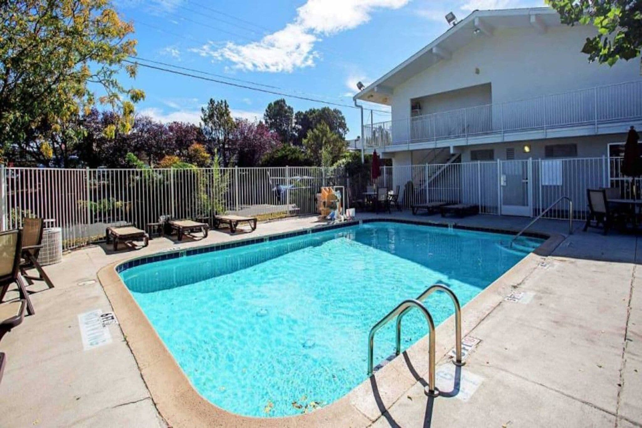 Une piscine rectangulaire avec un escalier en métal, entourée d’une clôture blanche, et un bâtiment avec un balcon en arrière-plan.