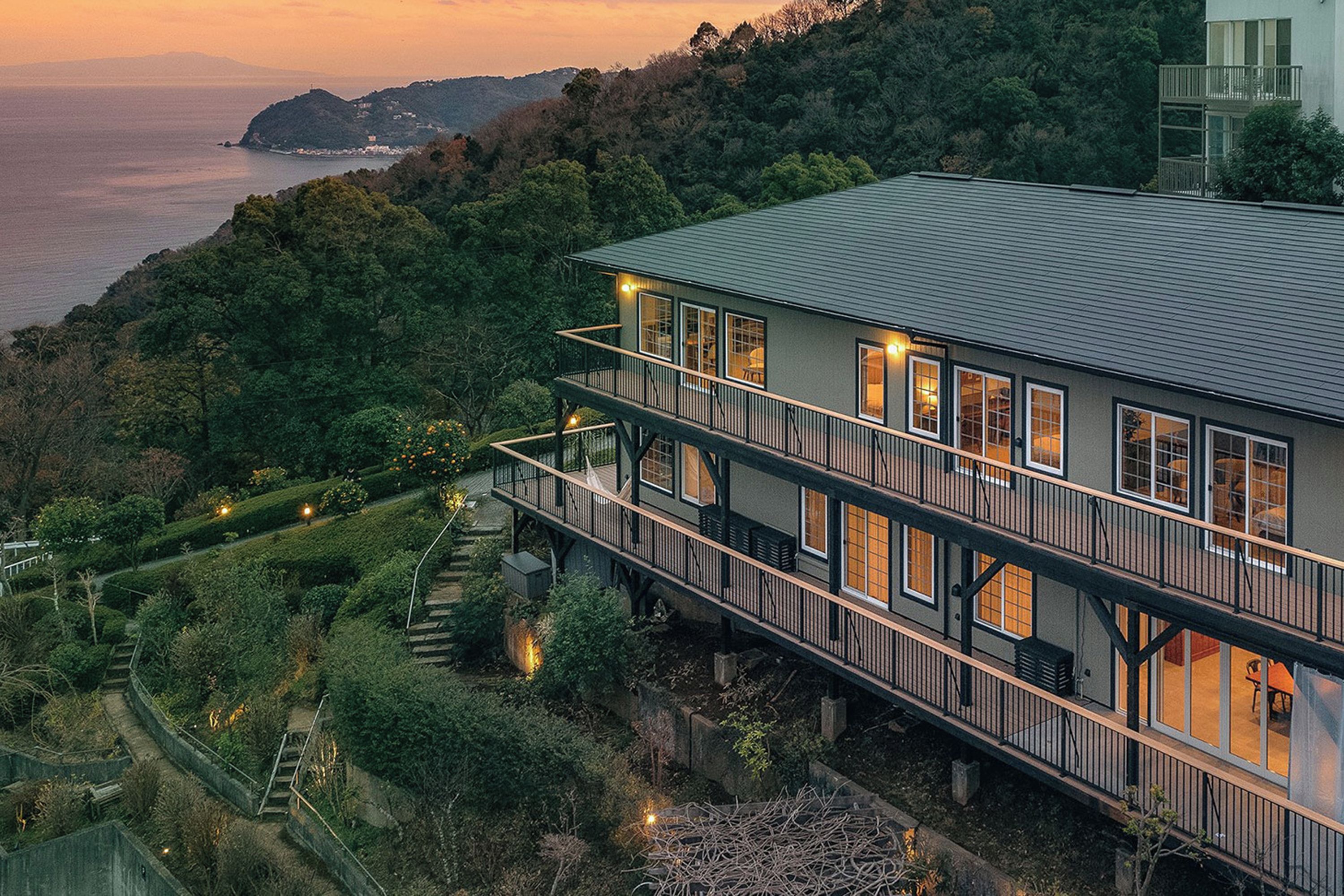 A multi-story building with a balcony, surrounded by lush greenery and overlooking a body of water.