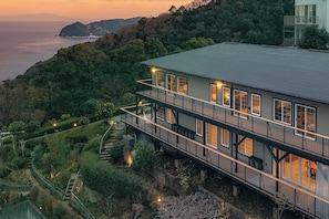 A multi-story building with a balcony, surrounded by lush greenery and overlooking a body of water. - GLAMDAY ATAMI (Atami)
