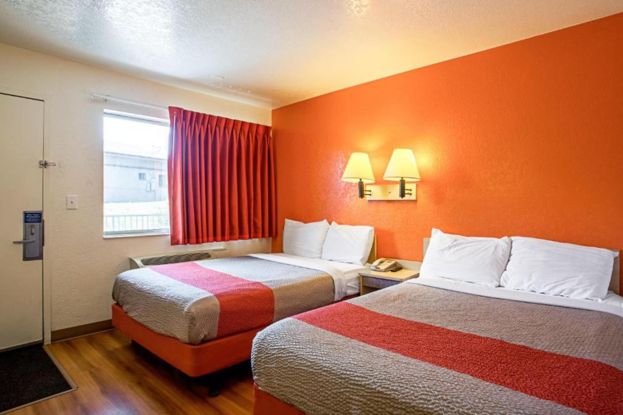 Une chambre d’hôtel avec deux lits, une fenêtre avec des rideaux, une lampe fixée au mur et un téléphone sur la table de chevet.