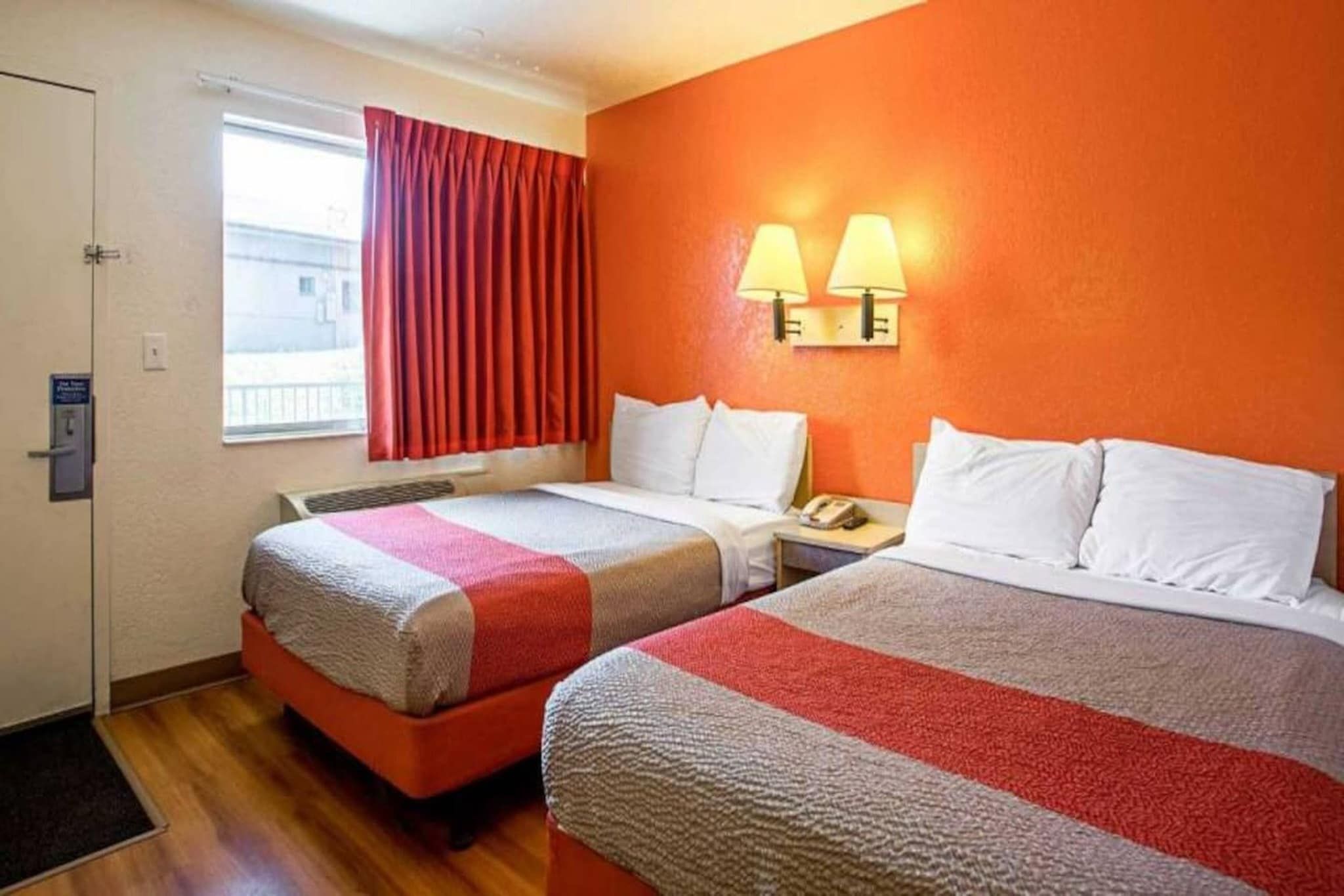 A hotel room with two beds, a window with red curtains, and a wall-mounted lamp.