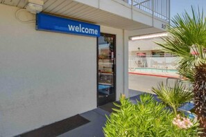 Entrada de un edificio con un letrero de 'bienvenida', una puerta de vidrio y una piscina visible a través del vidrio.