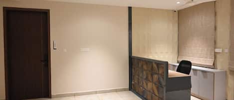 A hotel room with a wooden door, a reception desk, a ceiling fan, and a window with a blind.