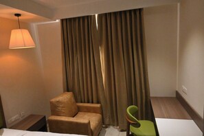 Habitación de hotel con un sillón de cuero marrón, una silla verde, un ventanal con cortinas marrones y una lámpara fijada a la pared.