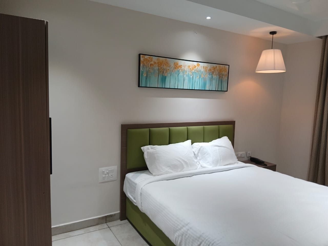 A hotel room with a bed, a painting on the wall, and a hanging light fixture.