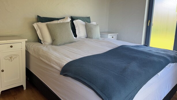 Ein Schlafzimmer mit einem Bett, weißen Nachttischen, einer Kommode und Blick durch eine Tür.