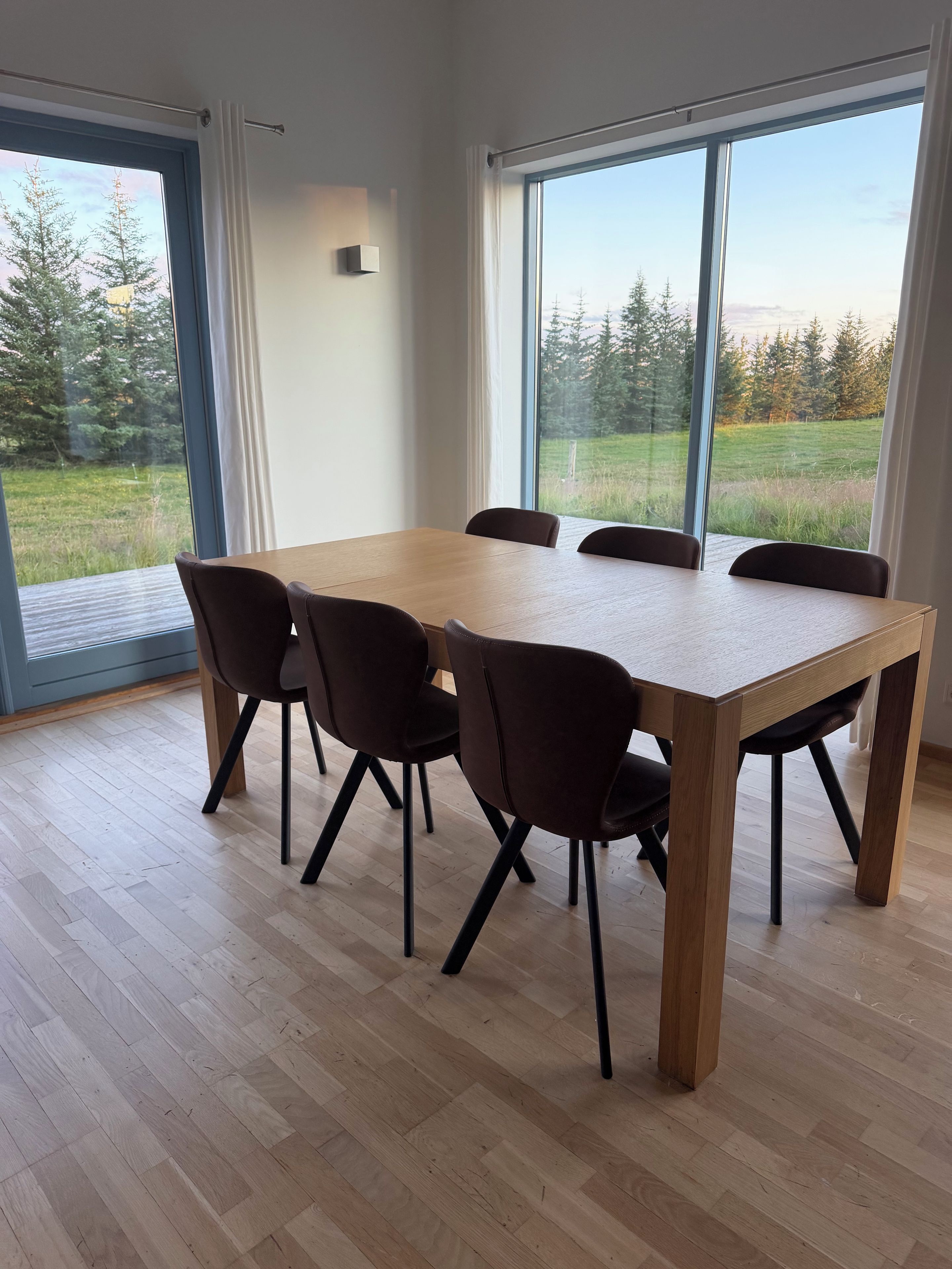 Een eethoek met een houten tafel en stoelen, grote ramen met uitzicht op bomen en gras.