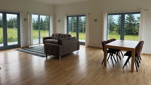 Ein geräumiges Zimmer mit Holzboden, einer Couch, einem Esstisch mit Stühlen und großen Schiebeglastüren, die zu einer grünen Außenfläche führen.