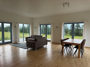 A spacious room with a wooden floor, a sofa, a dining table with chairs, and large sliding glass doors leading to a grassy outdoor area. - Marvelous 3-bedroom farmhouse in charming Rangárþing ytra south Iceland (Rangárþing ytra)