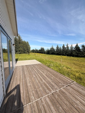 A wooden deck leading to a glass door, surrounded by a grassy field and a line of trees.