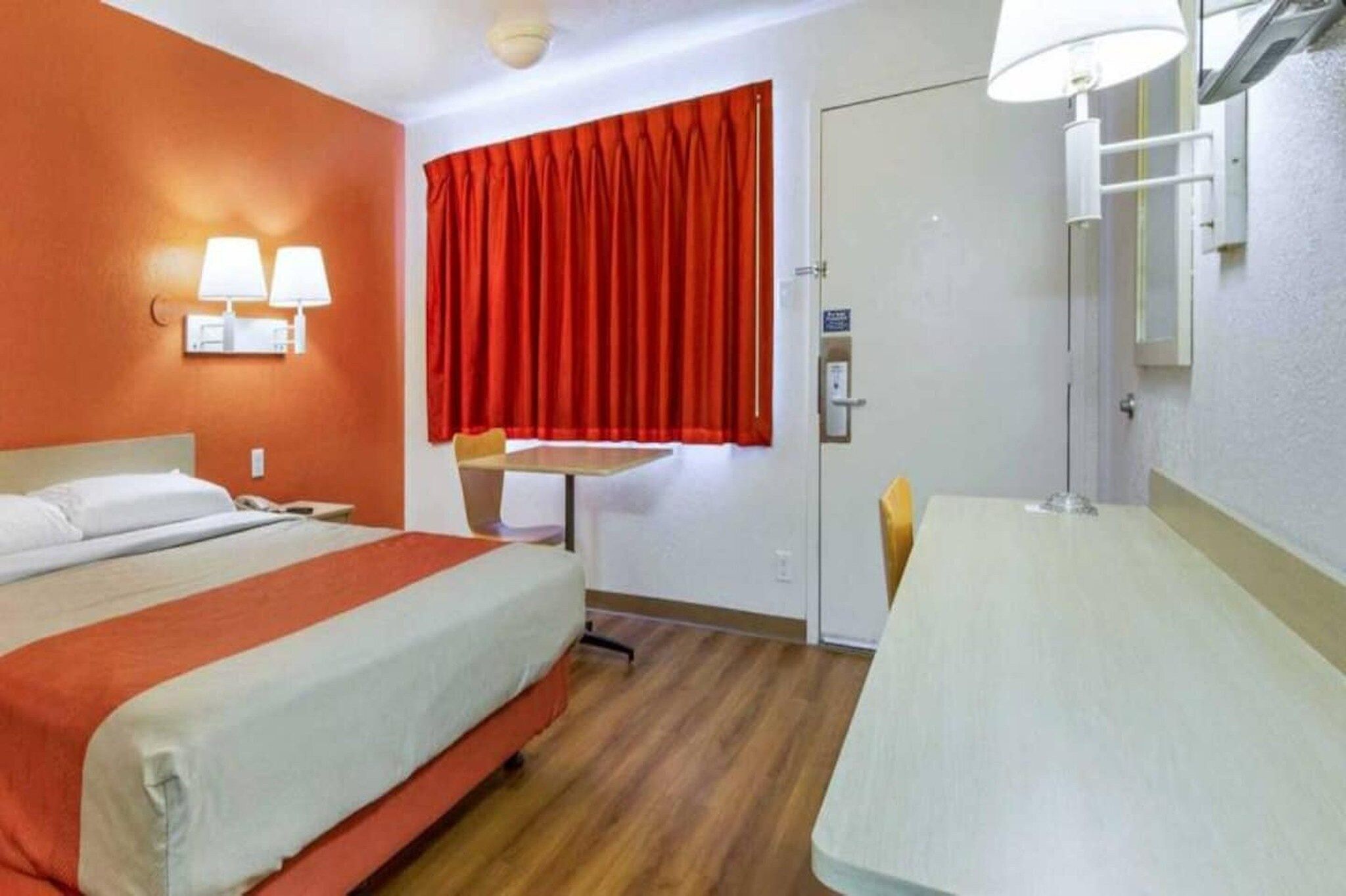 Habitación de hotel con cama, escritorio, silla, ventana con cortinas rojas y lámpara de pared.