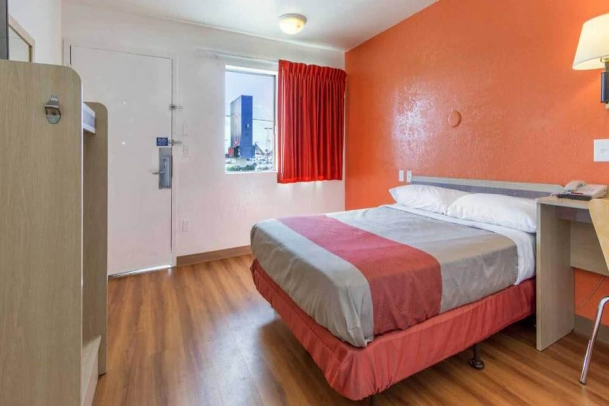 A hotel room with a bed, a desk, and a window with red curtains.