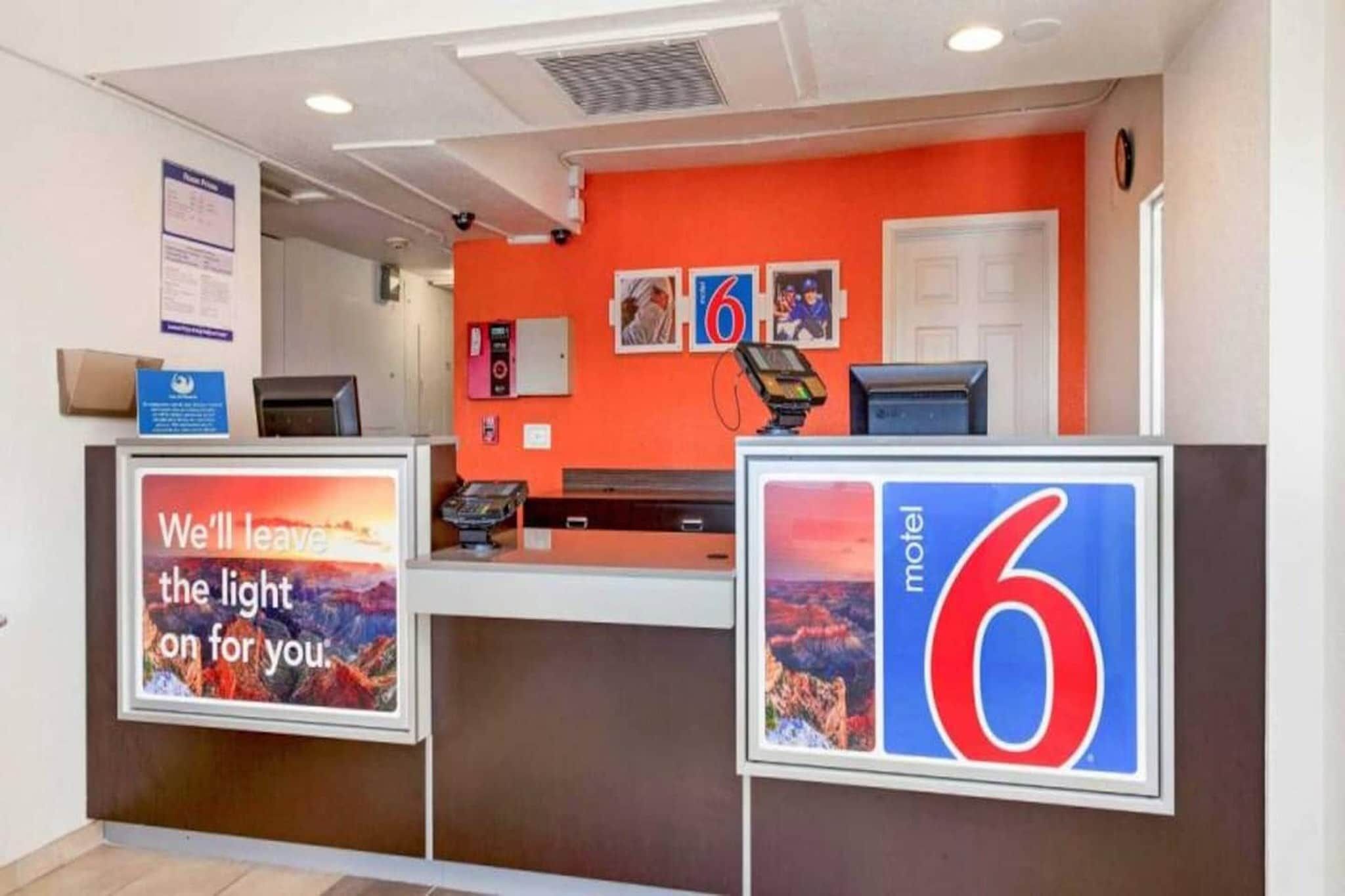A hotel reception area with a red wall, a number '6' sign, and a sign that says 'We'll leave the light on for you.'