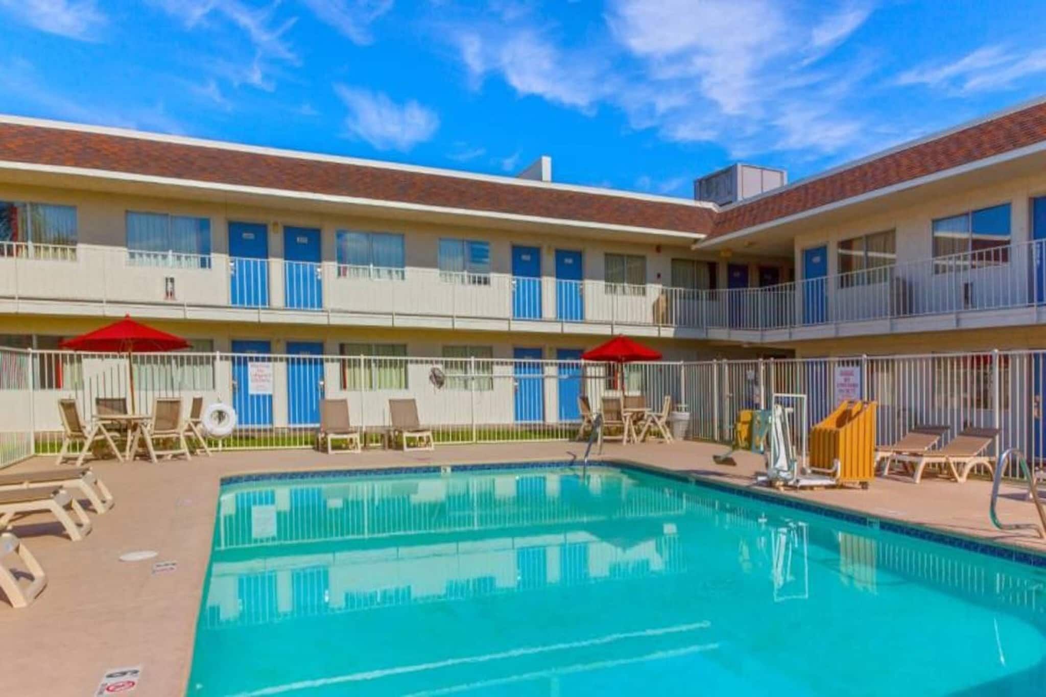 Una piscina con sillones y sombrillas, rodeada por un edificio de varios pisos con balcones.