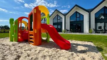 A playground with a red slide and climbing structures, located in front of a modern building with large windows.