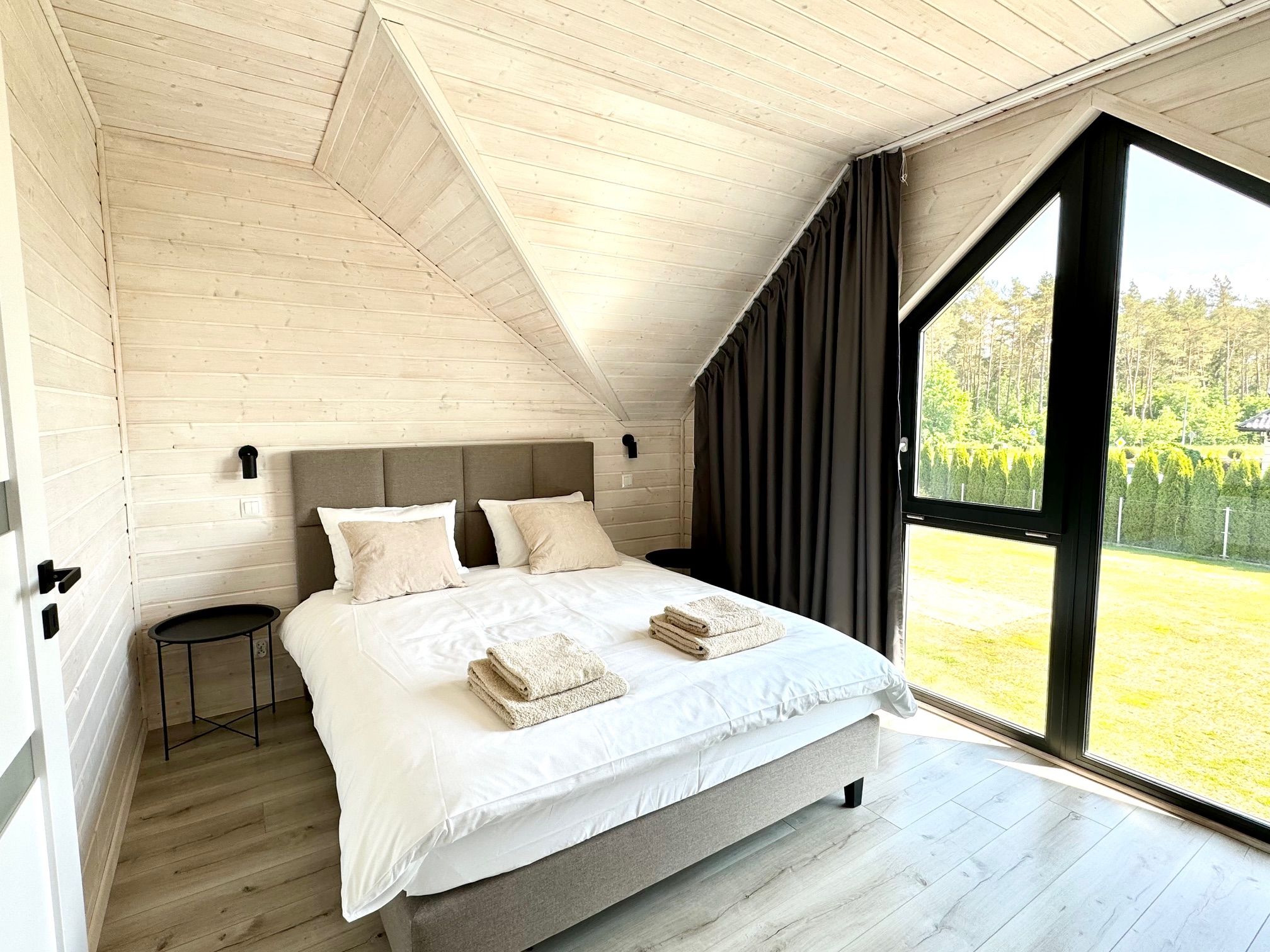 A bedroom with a large bed, a small table, and a window with a view of greenery.