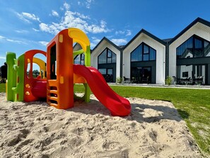 A playground with a slide and climbing structures, set against a modern building with large windows.