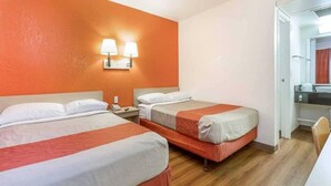 Quarto de hotel com duas camas, uma mesa de cabeceira, uma luminária e um banheiro visível por uma porta aberta.