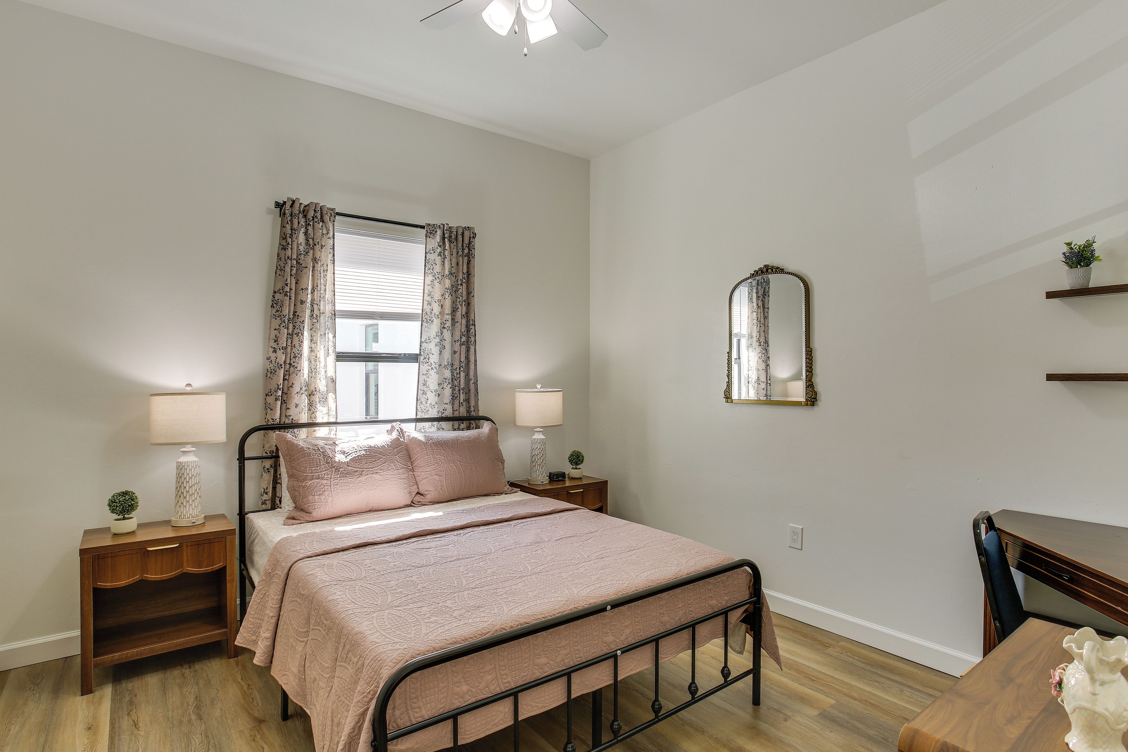 Quarto com cama, duas mesas de cabeceira, um criado-mudo, espelho, janela com cortinas e cômoda de madeira.