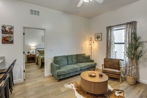 A living room with a green sofa, a round wooden coffee table, a small side table, a floor lamp, a chair, and a large window with curtains. - Stylish Condo: Walk to Caloosahatchee River (LaBelle)