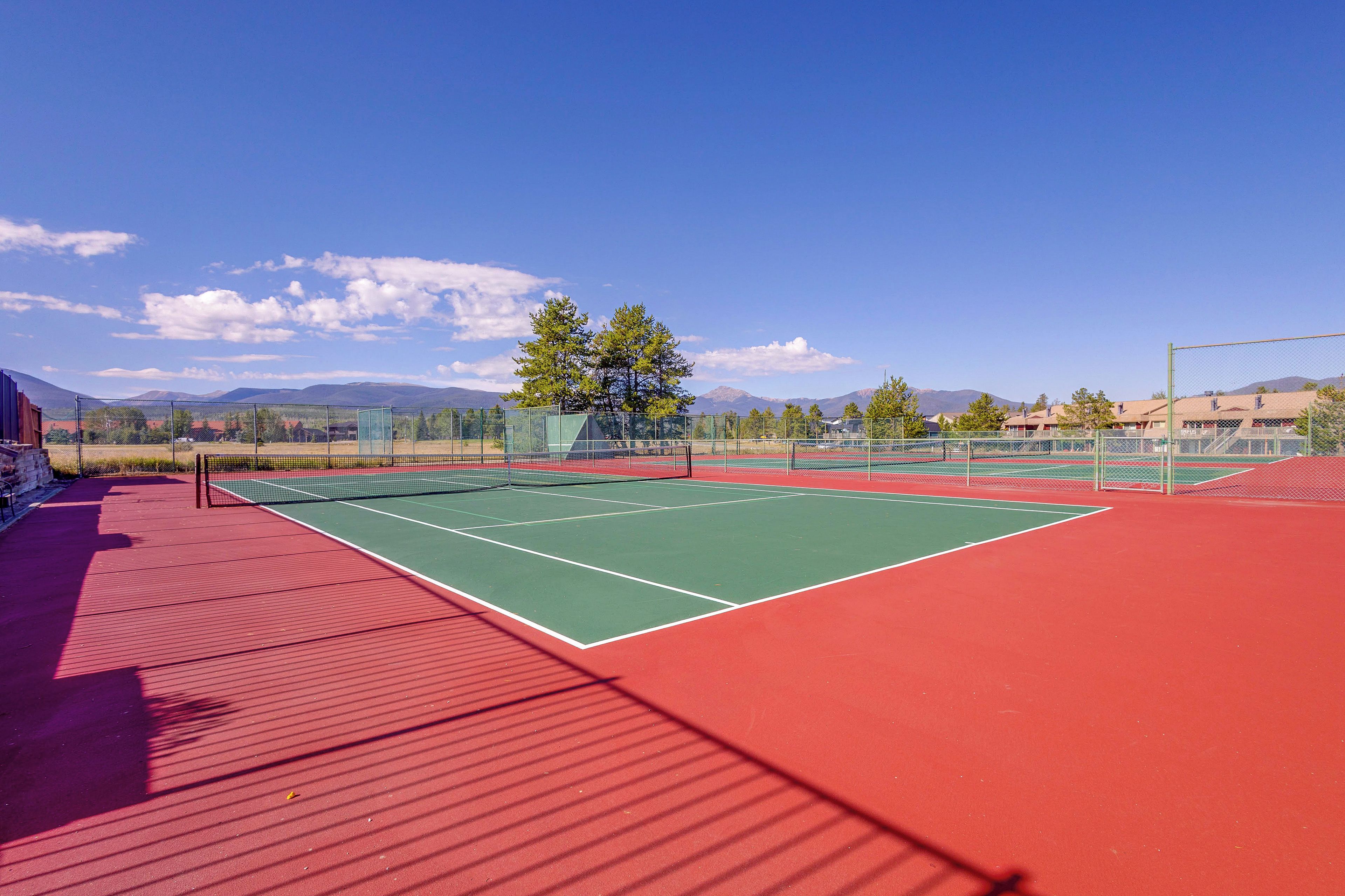 Ein Tennisplatz im Freien mit roter Belag und grünen Spielfeldern, umgeben von einem Maschendrahtzaun, mit Bergen im Hintergrund.