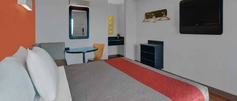 Habitación de hotel con cama, escritorio, silla, mesita, espejo, televisor y armario.