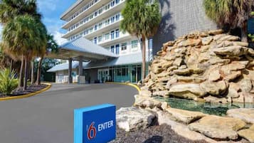 Entrada de un hotel con un letrero azul que dice '6 ENTER', una zona de estacionamiento techada y una formación rocosa con agua.