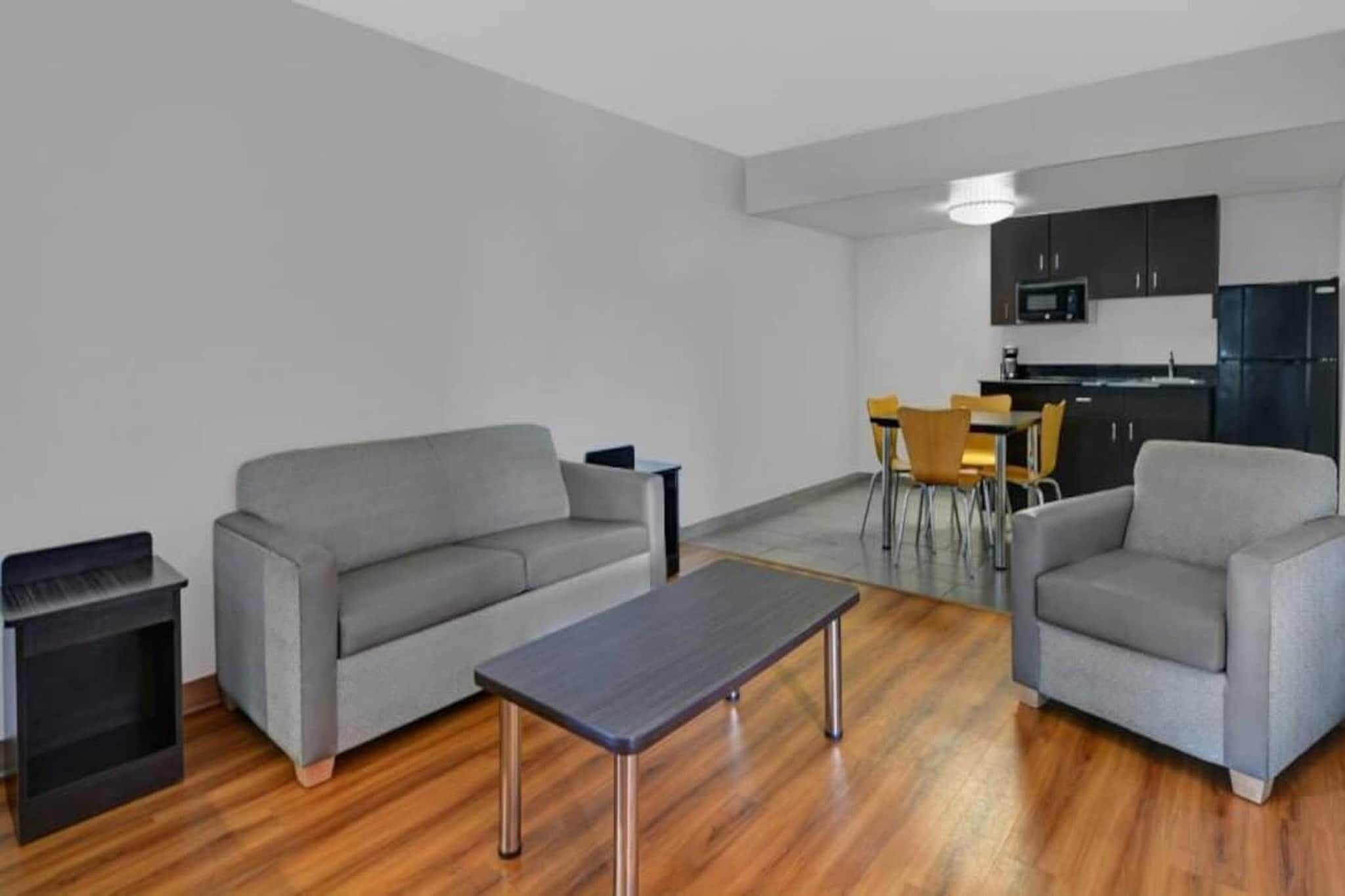 A modern living room with a grey sofa, a coffee table, and a dining area with chairs and a kitchen in the background.