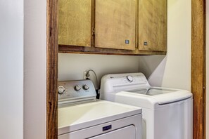 A compact laundry area with a washing machine and dryer, wooden cabinets above, and a white wall. - Fire Pit, Beautiful Wooded View, 70 Huntable Acres (Mecosta)