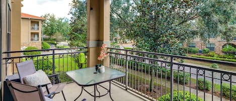 A balcony with a glass table and chairs, a flower vase, and a view of a landscaped garden and buildings.