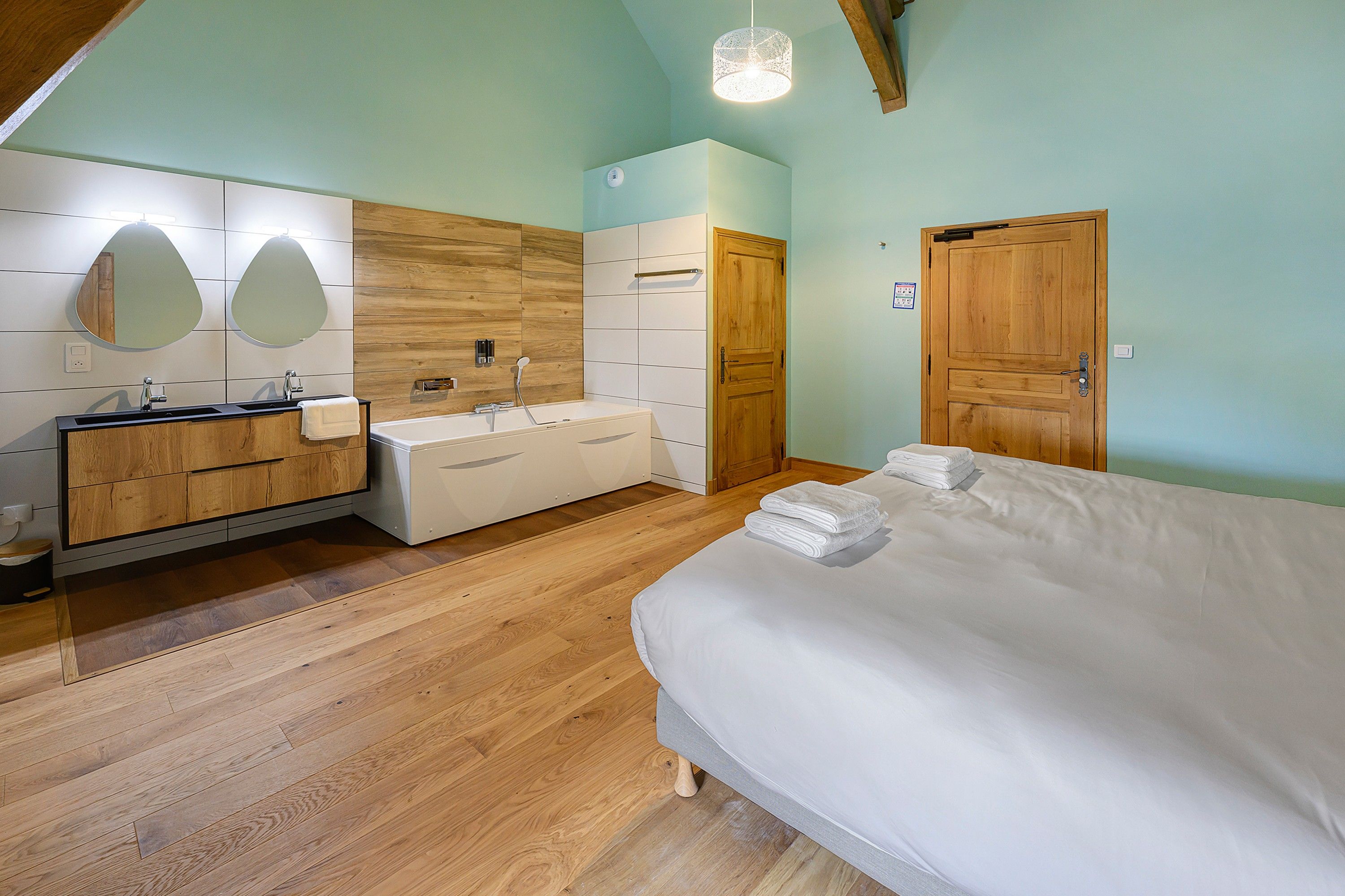 A bedroom with a wooden floor, a bed with white linens, a wooden wardrobe, a bathroom with a mirror, and a wooden door.