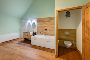 Ein modernes Badezimmer mit Holzboden, einer weißen Badewanne, einem Waschbecken mit Spiegel und einer Toilette.