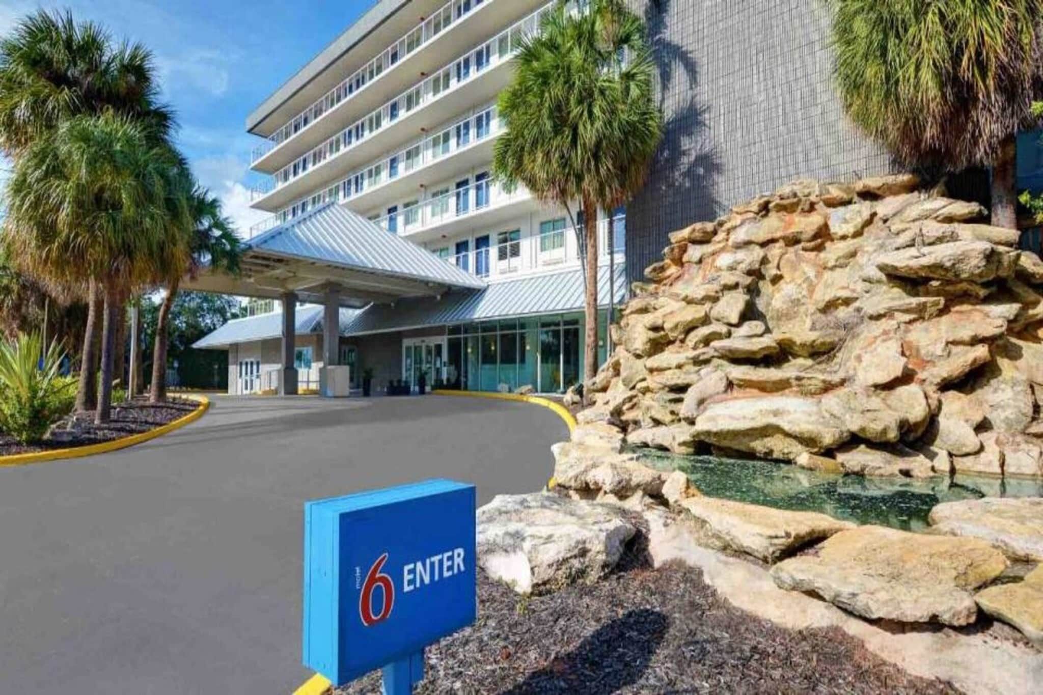 A hotel entrance with a blue sign indicating the entrance, a covered parking area, and a rock formation with a water feature.