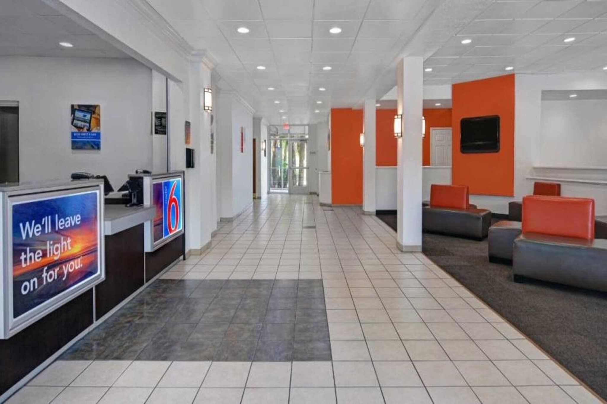 A hotel lobby with a reception desk, a sign reading 'We'll leave the light on for you.', and a seating area with orange and grey furniture.