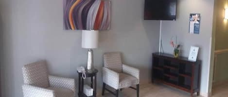 A hotel lobby with two patterned armchairs, a small table with a lamp, a television on the wall, and a reception desk.