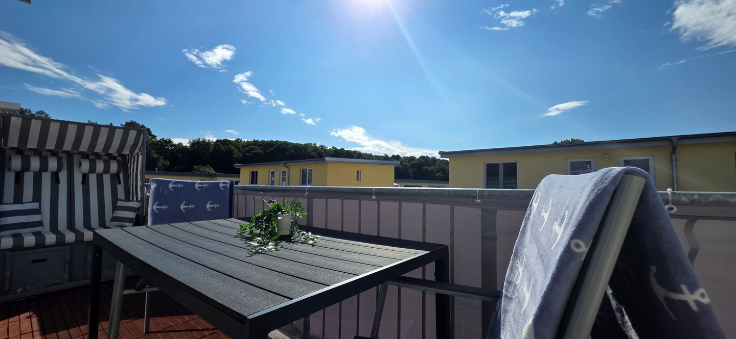 Eine Terrasse im Freien mit Tisch und Stühlen, einer kleinen Pflanze und Blick auf ein gelbes Gebäude im Hintergrund.