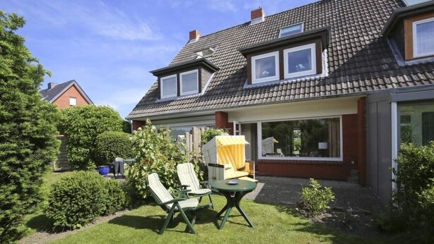 Ein Wohnhaus mit Satteldach, zwei Fenstern, einer überdachten Außenveranda mit Stühlen und Tisch und einem gepflegten Rasen.
