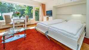 Ein Hotelzimmer mit Bett, Fernseher, Essbereich mit Tisch und Stühlen sowie Blick auf Grünflächen.