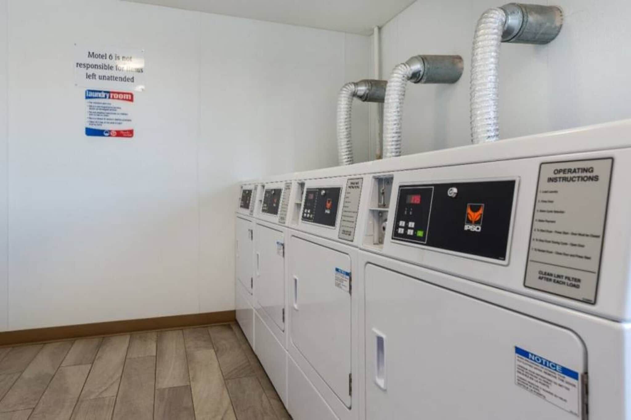 A row of white laundry machines with control panels, a notice about unattended luggage, and a sign for operating instructions.