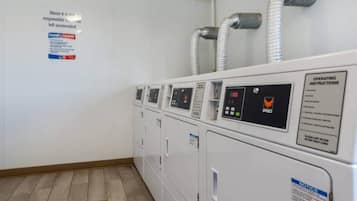 A row of white laundry machines with control panels, a notice about unattended luggage, and a sign for operating instructions.