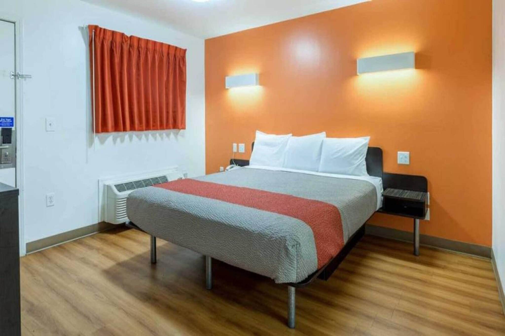 A hotel room with a bed, a heater, a window with curtains, and a wall-mounted light fixture.